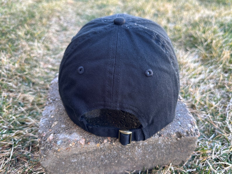 K&ouml;nnte beinhalten: Schwarze Baseballkappe mit einem schwarzen verstellbaren Riemen und zwei kleinen schwarzen &Ouml;sen auf der R&uuml;ckseite.