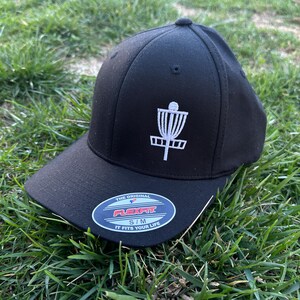 May include: Black baseball cap with a white embroidered disc golf basket design. The cap has a Flexfit label with the text "The Original Flexfit S/M It Fits Your Life."