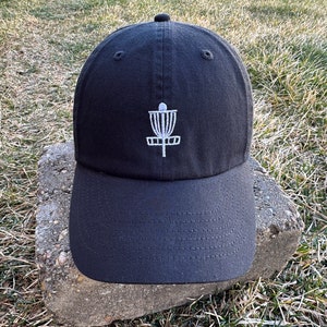 K&ouml;nnte beinhalten: Eine schwarze Baseballkappe mit einem wei&szlig;en gestickten Disc-Golf-Korb-Design.