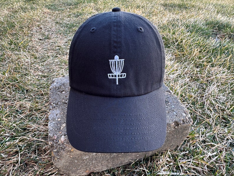 K&ouml;nnte beinhalten: Eine schwarze Baseballkappe mit einem wei&szlig;en gestickten Disc-Golf-Korb-Design.