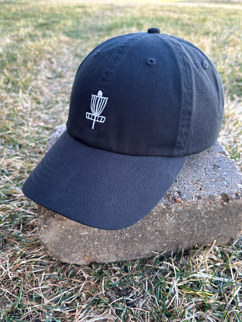 K&ouml;nnte beinhalten: Eine schwarze Baseballkappe mit einem wei&szlig;en gestickten Disc-Golf-Korb-Design.