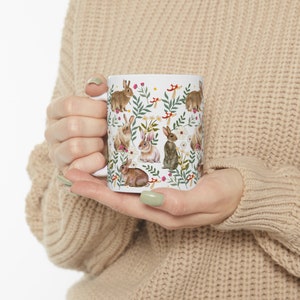 May include: White ceramic mug with a repeating pattern of brown rabbits, green leaves, white flowers, and red mushrooms.