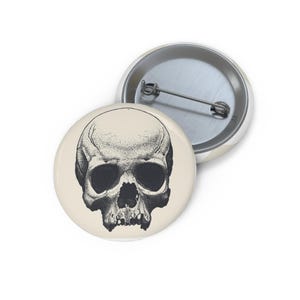 May include: A round pinback button featuring a detailed black and white stippled illustration of a human skull centered on a cream-colored background. The back of the button is metallic silver and includes a horizontal pin fastener.
