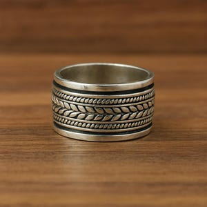 Puede incluir: Un anillo ancho de color plateado con un patrón detallado. El anillo presenta una serie de bandas texturizadas, incluyendo una banda central con un diseño en forma de hoja. El anillo se muestra sobre una superficie de madera.