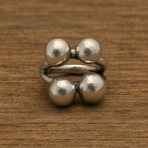 May include: A silver-colored ring featuring four spherical accents. The ring has a unique design with the spheres arranged in two pairs, connected by a curved band. The ring is displayed on a wooden surface.