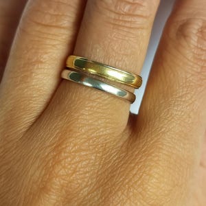 May include: A close-up shot of two rings, one gold and one silver, worn on a finger. The gold ring sits above the silver one. Both rings have a plain, rounded design. The background is blurred skin.