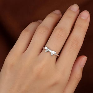 May include: A silver ring with a bow-shaped design. The bow is the focal point, positioned centrally on the ring's band. The ring's design is minimalist and elegant. The ring is shown on a finger, showcasing its size and style.