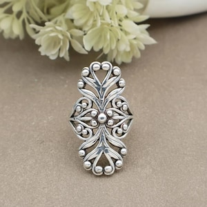 May include: A silver-toned ring with an ornate, symmetrical design. The ring features a central floral-like motif surrounded by stylized leaves and small, round accents. The ring is set against a neutral background, with white flowers in the background.