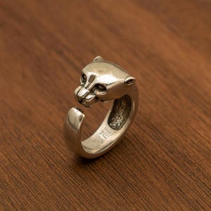 May include: A silver-toned ring featuring a detailed panther head design. The ring's head is open, creating a unique wrap-around style. The ring is set against a warm, wooden background, highlighting its metallic sheen.