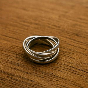 Puede incluir: Un anillo entrelazado de plata con cinco bandas. Las bandas son lisas y brillantes, creando un diseño moderno y elegante. El anillo está sobre una superficie de madera marrón cálida.
