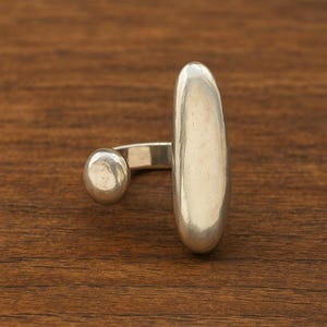 Puede incluir: Un anillo de plata con una forma ovalada alargada y un pequeño acento redondo. El anillo se encuentra sobre una superficie de madera marrón, mostrando su acabado metálico pulido. El diseño es moderno y minimalista.
