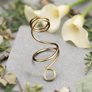 May include: A gold-toned metal ring featuring a continuous spiral design that wraps around the finger. The wire structure forms multiple circular loops of varying sizes, ending in a small, rounded terminal point at the top and bottom.