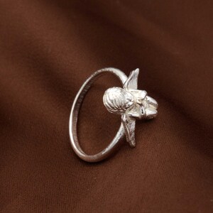 May include: A silver ring featuring a detailed floral design. The ring has a circular band with a sculpted flower and leaf motif. The ring is made of a shiny, metallic material and is set against a brown background.