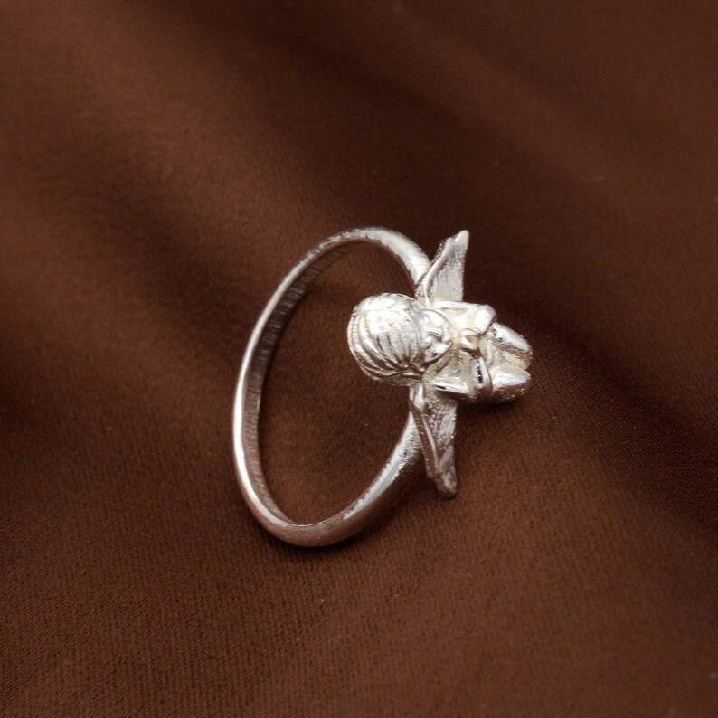 May include: A silver ring featuring a detailed floral design. The ring has a circular band with a sculpted flower and leaf motif. The ring is made of a shiny, metallic material and is set against a brown background.
