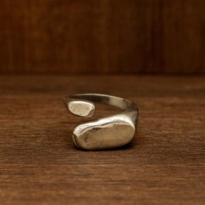 May include: A silver ring with an open design. The ring has two rounded, smooth, and slightly flattened oval shapes. The ring is set against a dark wood grain background.