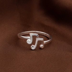 May include: A silver-colored ring featuring two musical notes. The ring is designed with a simple band and the notes are positioned side by side. The ring is made of a shiny, metallic material.