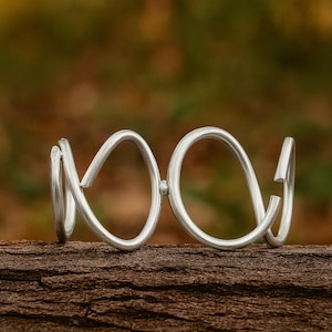 May include: A silver-colored bracelet with a unique, open design. The bracelet features a series of interconnected, looped shapes, creating an abstract and modern aesthetic. The jewelry is displayed on a piece of wood, with a blurred natural background.
