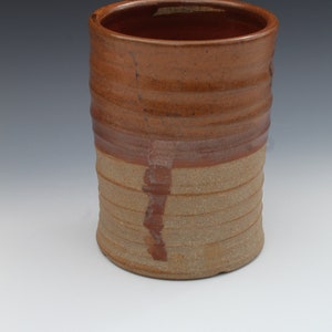 Puede incluir: Una taza de cerámica marrón con un esmalte marrón claro y un esmalte marrón oscuro en la mitad superior. La taza tiene una superficie texturizada y una forma ligeramente redondeada.
