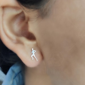 Puede incluir: Un pendiente de plata con forma de figura corriendo se ve en una oreja. El diseño del pendiente es minimalista, representando a una persona en movimiento. El fondo está borroso, destacando la joya.