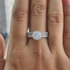 Puede incluir: Un juego de anillos de tres piezas con una gema grande, redonda y transparente y piedras más pequeñas. El anillo principal tiene un halo de piedras más pequeñas alrededor de la piedra central. Los otros dos anillos tienen piedras más pequeñas. Los anillos están engastados en una banda plateada.