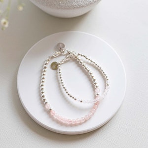 May include: Two bracelets, one with silver beads and one with pink beads, on a white surface.