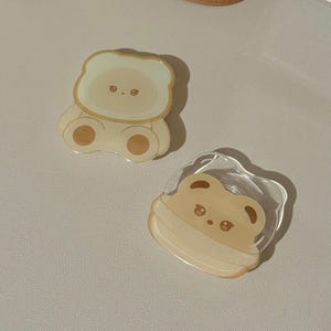 May include: Two small, clear resin bear charms with brown eyes and a light brown outline. The bear on the left is wearing a light green hooded jacket. The bear on the right is wearing a light brown scarf.