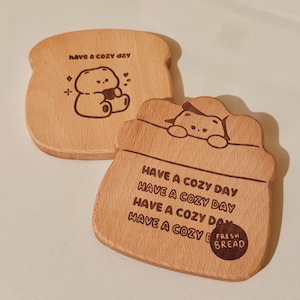 May include: Two wooden coasters shaped like bread slices. One coaster features a cartoon bear and the text "have a cozy day." The other coaster has the text "Have a cozy day" repeated and "Fresh Bread."