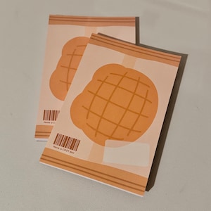 May include: Two beige notepads with a cartoon illustration of a melon pan. The melon pan is orange with a grid pattern. The notepads have a barcode and the text "Have a COZY day".