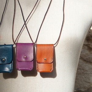May include: Three leather pouches with different coloured straps hanging from a white mannequin. The pouches are teal, purple, and brown.