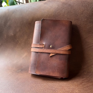 Op de afbeelding: Een handgemaakt, donkerbruin leren notitieboek met een wikkelriemsluiting. Het notitieboek is gesloten, met een zichtbare messing knop. Het leer heeft een natuurlijke, getextureerde uitstraling. Het notitieboek is een klassiek, tijdloos ontwerp.