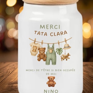 May include: A white ceramic jar with a wooden lid featuring a printed design of a clothesline with a baby onesie, teddy bear, overalls, leaf, hat, and airplane. The text reads MERCI TATA CLARA, MERCI DE T'ÊTRE SI BIEN OCCUPÉE DE MOI, and NINO.