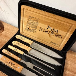 Peut inclure: Un étui noir zippé contenant une planche à découper en bois et des ustensiles de barbecue. La planche est gravée d'une illustration de grill et du texte "Papa LE CHEF DU GRILL" et "Angélica". L'ensemble comprend des couteaux et une fourchette.