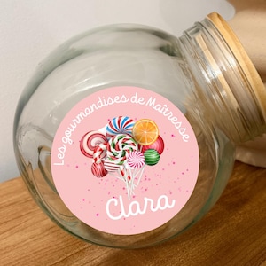 Peut inclure: Un bocal en verre transparent avec un couvercle en bois, orné d'une étiquette rose avec des sucettes colorées et le nom "Clara". L'étiquette comprend également le texte "Les gourmandises de Maîtresse".