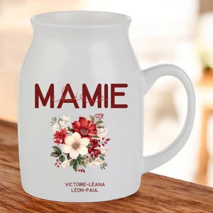 Op de afbeelding: Witte keramische mok met een handvat, met het woord "MAMIE" in bordeauxrode letters boven een bloemstuk van rode en witte bloemen. De namen "VICTOIRE-LÉANA" en "LÉON-PAUL" staan eronder. De mok heeft een gebogen vorm.