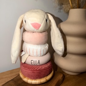 Peut inclure: Un jouet d'empilage en forme de lapin en peluche, avec une tête crème, un nez rose et de longues oreilles. Le jouet comporte plusieurs anneaux aux couleurs pastel, dont un avec le nom "ELLA" imprimé. La base est en bois.