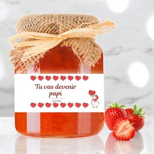 Puede incluir: Un tarro de cristal lleno de mermelada roja, sellado con una tapa de arpillera y atado con un lazo de rafia. Una etiqueta blanca dice "Tu vas devenir papi" con gráficos de fresas y corazones. Fresas frescas delante del tarro.