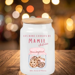 Peut inclure: Un pot à biscuits en céramique blanche avec un couvercle en bois. Le pot porte l'inscription "LES BONS COOKIES DE MAMIE chérie" et une illustration de biscuits dans un bocal. Les noms "lola, lucas & milana" sont imprimés en bas.