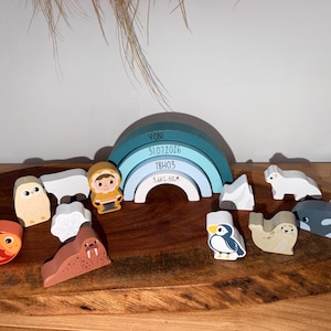 Peut inclure: Un ensemble de figurines en bois sur le thème arctique, incluant un poisson, un hibou, un morse, un ours polaire, un personnage en parka, un phoque, un manchot et une baleine. Un arc-en-ciel bleu central porte le texte YONI, 31.07.2026, 18H03, 3.6KG-51CM.
