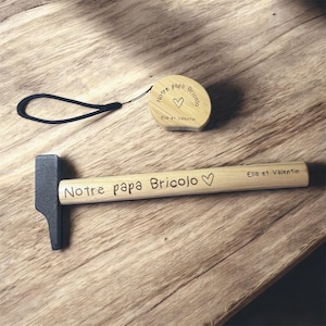 May include: A wooden hammer and a round wooden tape measure with black text and a heart symbol. The hammer handle reads "Notre papa Bricolo" and the tape measure reads "Notre papa Bricolo" and "Elia et Valentin".