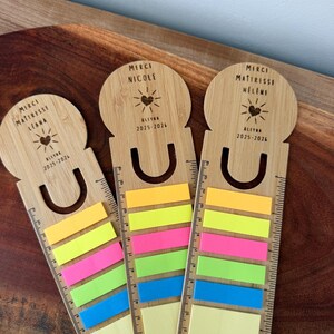 May include: Three light brown wooden bookmarks with integrated rulers and colourful sticky notes. Each bookmark features a circular top with engraved text including "MERCI MAITRESSE," a heart icon, and "ALEYNA 2025-2026." The rulers show markings up to 20 cm.