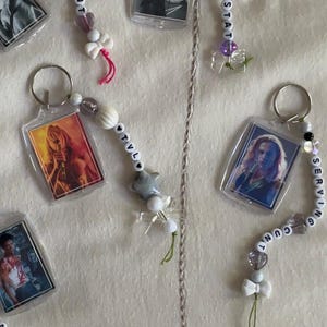 May include: Five rectangular acrylic keychains featuring character portraits. Each keychain has a metal ring and a beaded strand with letter beads spelling "SERVING CUNT," "LESTAT," "TVL," or "LOUSTAT," accented with small heart, bow, and star-shaped beads.