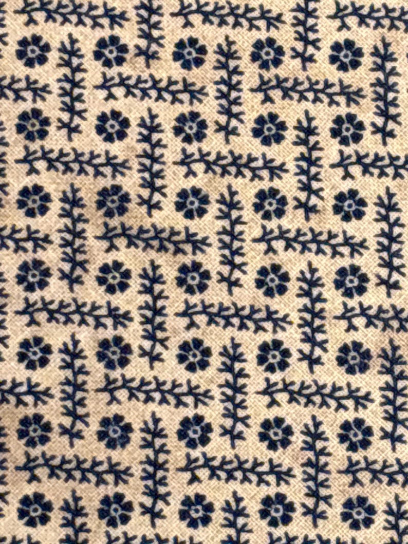 Puede incluir: Primer plano de una tela con un patr&oacute;n repetitivo de motivos florales y de hojas azul marino sobre un fondo beige. El dise&ntilde;o presenta peque&ntilde;as flores estilizadas y delicados elementos en forma de rama dispuestos en una formaci&oacute;n similar a una cuadr&iacute;cula.