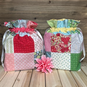 May include: Two patchwork drawstring bags with floral and geometric patterns in red, pink, green, and blue. Each bag has a ruffled top and white ribbon drawstring. A pink flower sits in front of the bags.