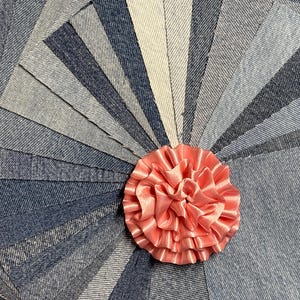 May include: A collection of rectangular denim fabric swatches in various shades of blue, light blue, and off-white, arranged in a fan pattern. A ruffled, circular peach-colored satin ribbon flower is positioned in the center of the fabric pieces.