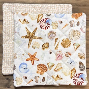 May include: Two quilted pot holders. The top pot holder features a white background with a pattern of seashells, starfish, and small blue starfish. The bottom pot holder has a beige floral pattern. The pot holders are square.