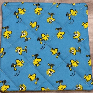 May include: Square, quilted pot holder with a light blue background and a repeating pattern of yellow cartoon birds. The pot holder has a yellow border and a diamond-shaped quilting pattern.