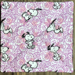 May include: Square quilted fabric featuring a repeating pattern of Snoopy in various poses, set against a backdrop of pink and white roses. The fabric is likely cotton or a similar material, suitable for quilting or crafting projects.