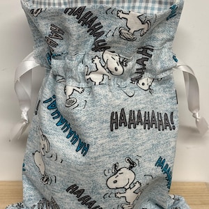 May include: A light blue drawstring bag featuring a repeating pattern of Snoopy characters and the word "HAHAHAHA!". The bag has a white ribbon drawstring closure and a gingham patterned interior.