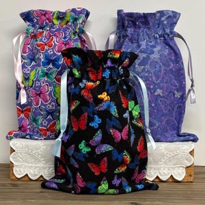 May include: Three drawstring bags with butterfly patterns. One bag is black with colorful butterflies, another is purple with a similar butterfly design, and the third is a lighter purple with a butterfly pattern. Each bag has a ribbon drawstring.