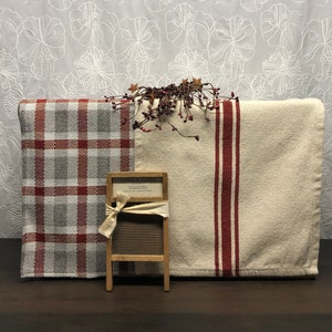 May include: A display featuring two folded towels, one with a red and gray plaid pattern and the other with a cream background and a red stripe. A miniature wooden washboard with a small sign and a decorative berry and star garland complete the arrangement.